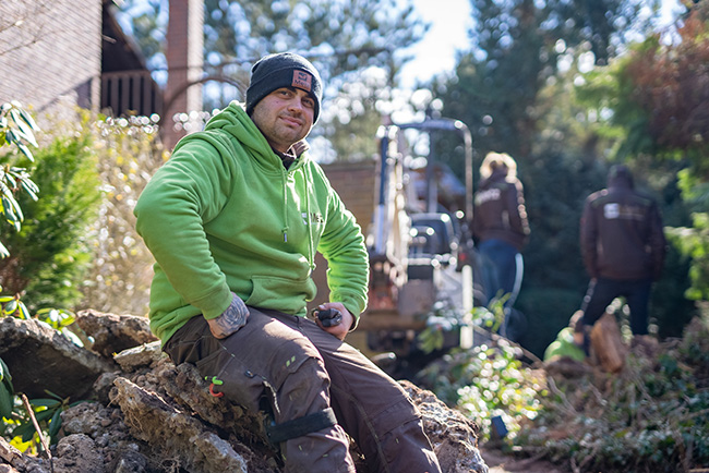 Timo sitzt in grüner Arbeitskleidung auf dem Boden