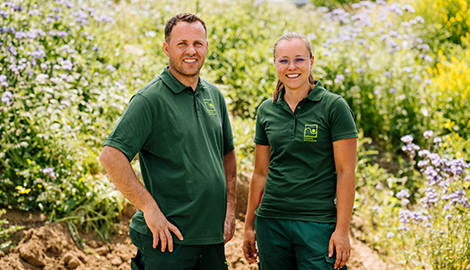 Ein Landschaftsgärtner und eine Landschaftsgärtnerin stehen vor einem begrünten Garten und lächeln in die Kamera