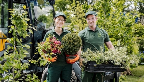 Zwei Landschaftsgärtner*innen stehen mit Pflanzen in der Hand vor der Kamera